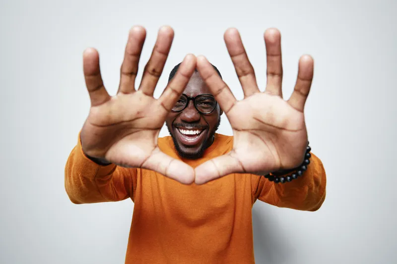 African American man with dark skin wearing glasses and orange sweater smiling and framing face with hands in heart shape on white background.