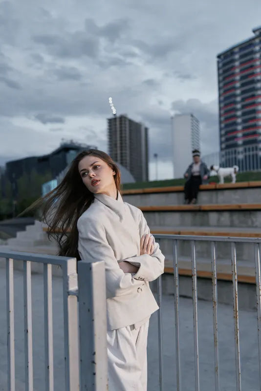 Woman in light coat standing outdoors with urban buildings and cloudy sky, casual confident expression, fashion style, overcast evening