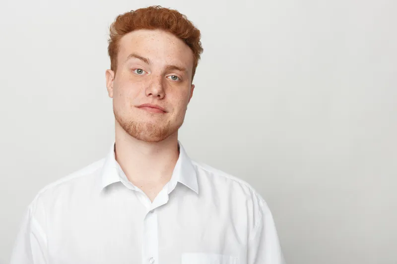 Young man with red hair and beard wearing white shirt looks confidently at camera on plain light background. Concept of casual style, portrait, and natural expression.
