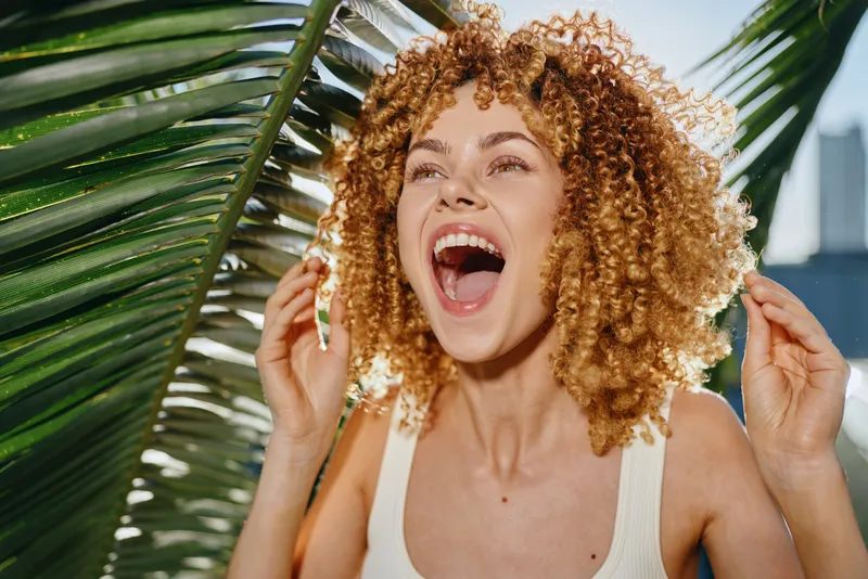 curly woman laughing joyfully outdoors near lush green palm leaves in bright sunlight, natural beauty and happiness captured in summer atmosphere