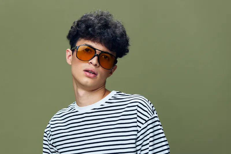 Young man with striped shirt and orange sunglasses posing against green background, showcasing casual style and curly hair in studio setting.