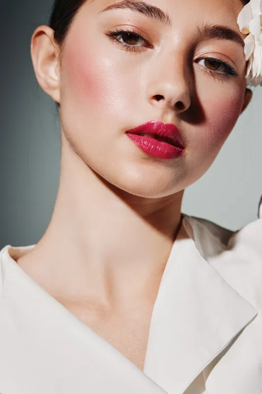 Close-up of a young woman with glossy skin wearing vibrant red lipstick and light makeup. Creative beauty and fashion concept with flattering studio lighting on gray background.