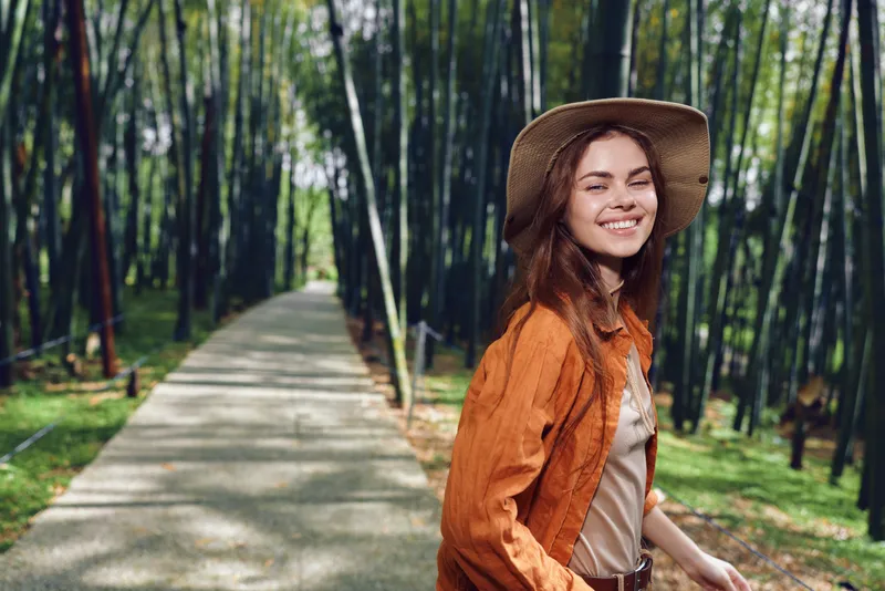 Woman smile hat bamboo path forest walking nature portrait of a young female traveler in a straw hat on a paved trail through a bamboo grove, casual orange shirt, joyful outdoor lifestyle.