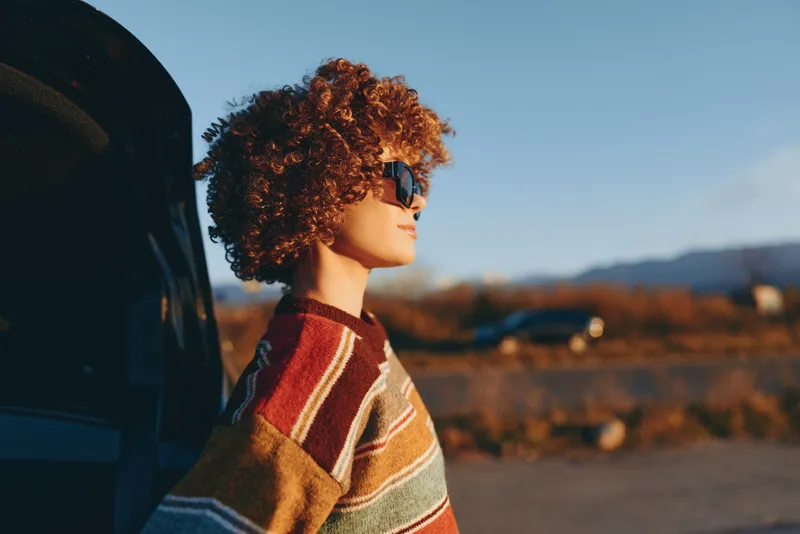 Woman with curly hair wearing a striped sweater outdoors near a car at sunset, portrait on a rural road, lifestyle moment in nature with warm natural light, calm and serene