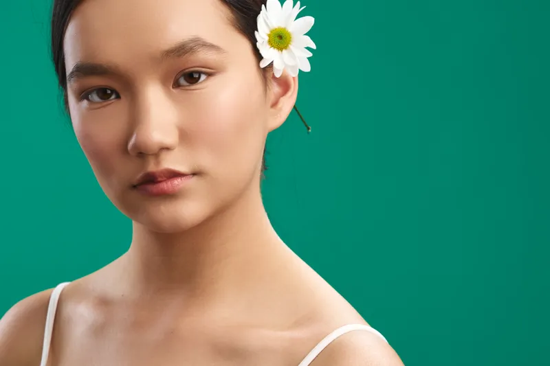 asian woman with a white flower tucked behind the ear stands before a vivid green backdrop, gentle expression and soft light creating a serene beauty portrait.