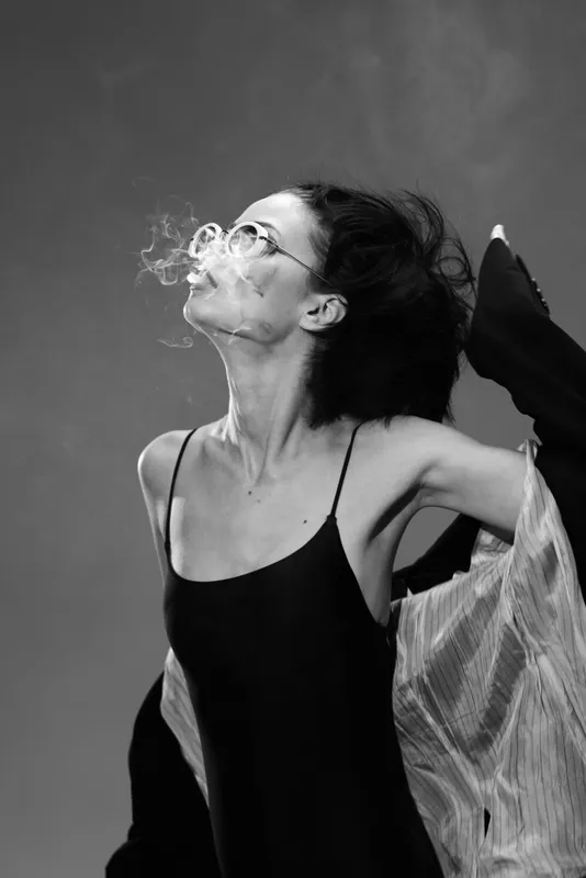Black and white portrait of a young woman with glasses blowing smoke, wearing a black dress and light outerwear in a studio setting.
