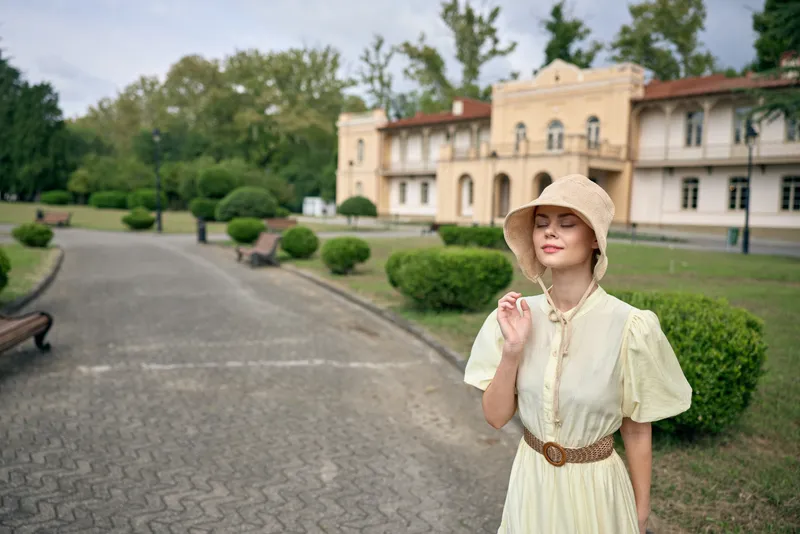 A woman in a pale yellow vintage dress with a wide belt and hat poses on a gravel path in front of a grand historic estate, smiling softly.
