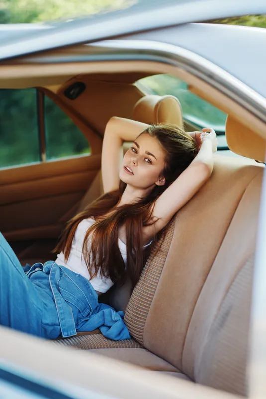Young woman relaxing in car interior with casual outfit, smiling and enjoying a sunny day, representing freedom and comfort during road trips.