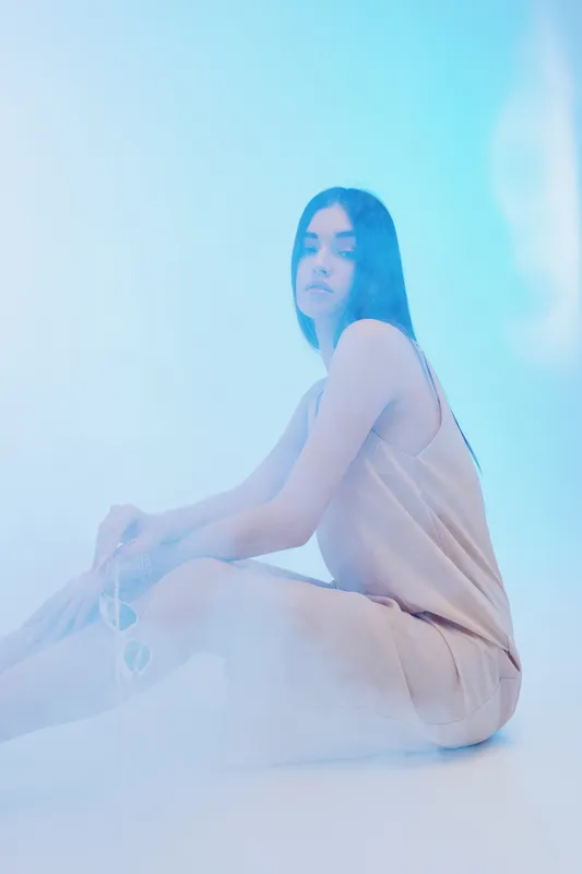 Young woman in beige dress sits on floor holding sunglasses in soft blue light. Minimalistic fashion portrait with serene expression and smooth skin. Concept of beauty and style.