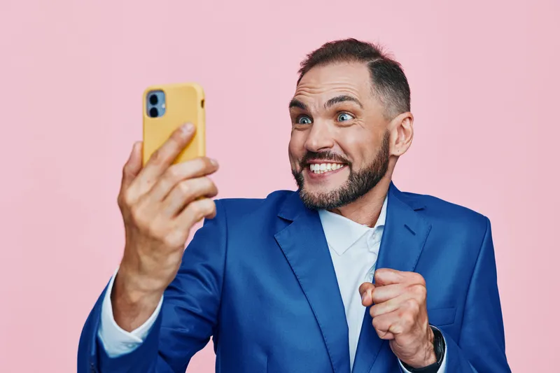 A professional man in a blue suit smiles passionately while taking a selfie on a vibrant pink background, illustrating confidence, energy, and positive momentum in a modern business moment.