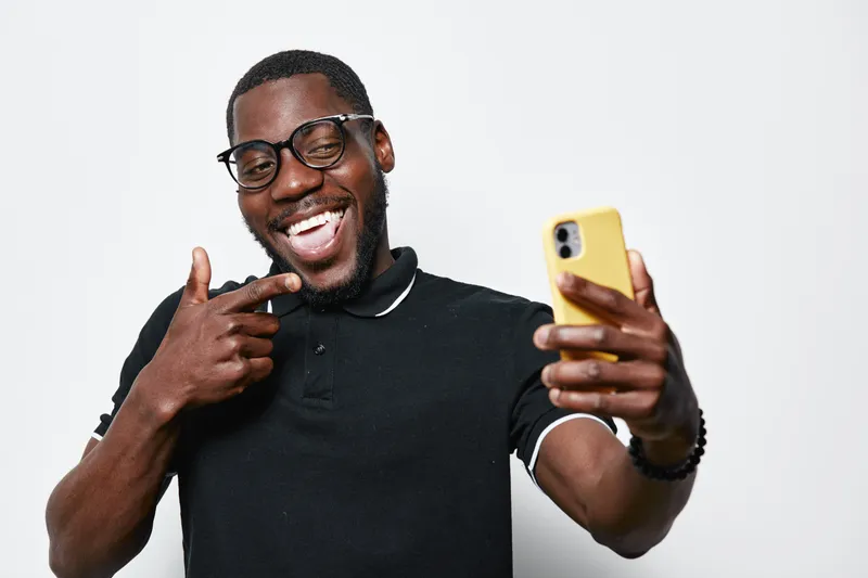 African American man with dark skin and glasses taking selfie with smartphone, smiling and pointing finger, casual black polo shirt on white background.