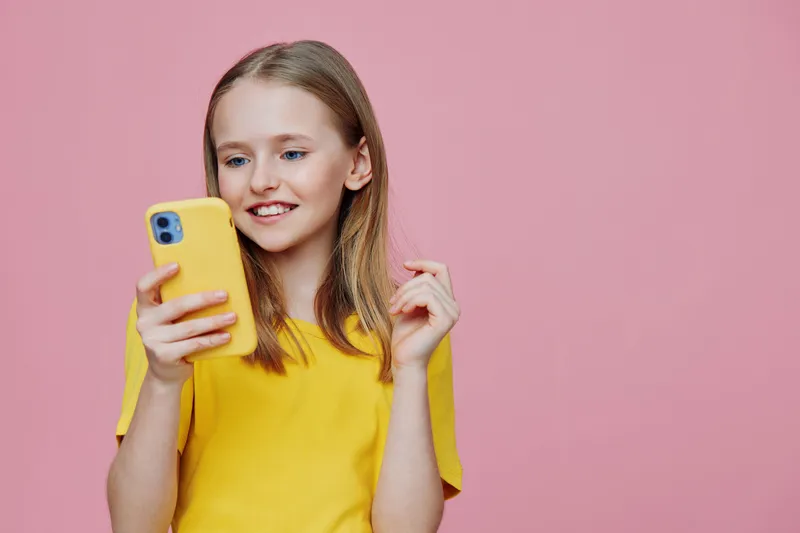 Happy girl in a yellow t shirt using a smartphone against a pink background, showcasing joy and connection through technology