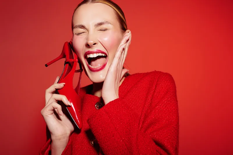 Happy woman in red sweater holds red high heel shoe near face, laughing with closed eyes on red background. Concept of fashion, joy, style, and vibrant emotions in studio.