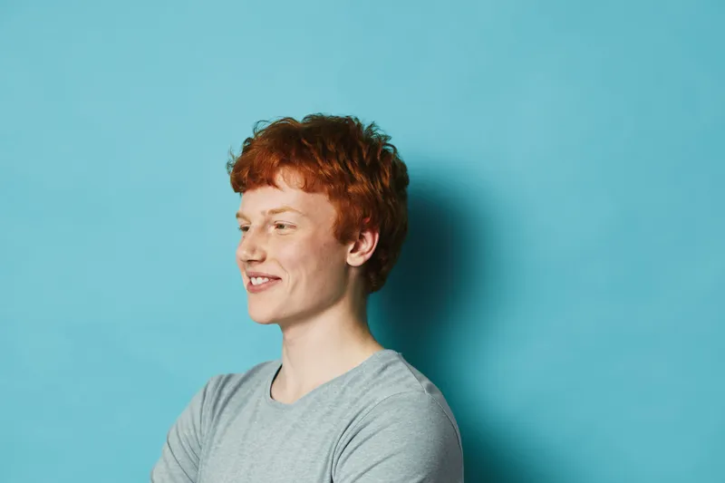 Portrait of young redhead man with curly hair and freckles, smiling in casual gray shirt against blue background, showing positive emotion and friendly expression.