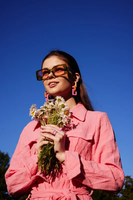 Young woman in stylish pink outfit holding daisies against a clear blue sky, showcasing summer fashion and vibrant colors