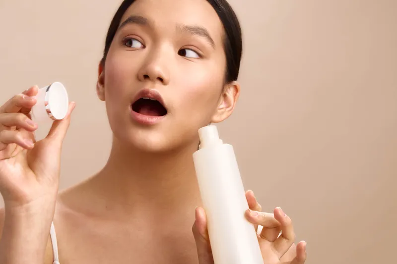 Asian woman poses in a bright setting, holding a lotion bottle and cap while applying skincare. Her expression is curious and engaged, creating a fresh, intimate beauty moment.