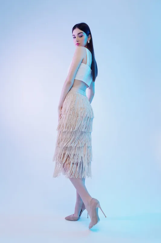 Elegant woman beauty fashion model poses in a fringed skirt and white top. Studio shot with soft blue lighting. Concept for style, glamour, and modern clothing trends.
