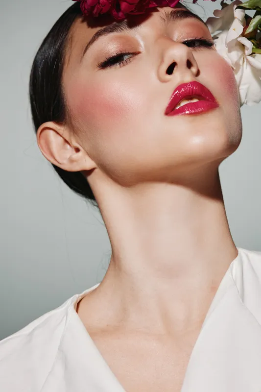 Close-up portrait of stylish woman with glossy red lipstick, flawless makeup, and floral arrangement in dark hair. Light background highlights edgy beauty and fashion concept.