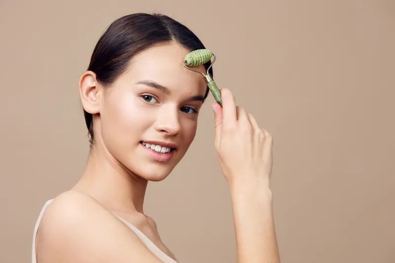 Young woman using a facial roller for skincare, promoting healthy beauty and wellness routines in a minimalist beige background