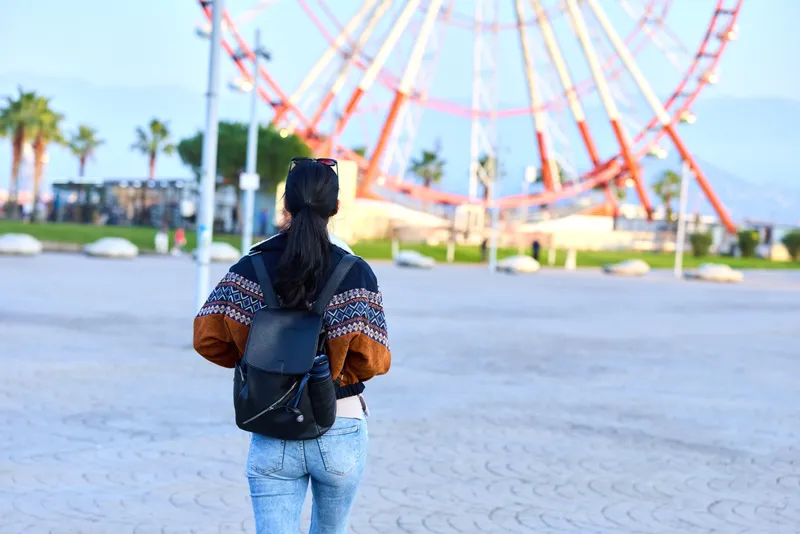 Traveler with backpack stands in a sunny amusement park, watching a towering rollercoaster, exploring outdoor scenery and casual fashion as distant rides spark curiosity