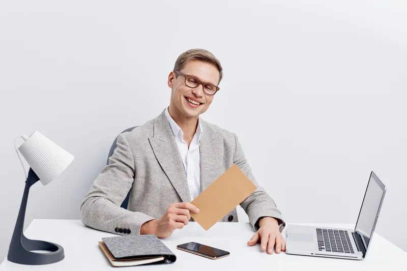 Smiling man in gray blazer holding envelope at desk with laptop, tablet, and smartphone, in modern office setting. Happy businessman with glasses working, positive mood, professional attire, bright ba
