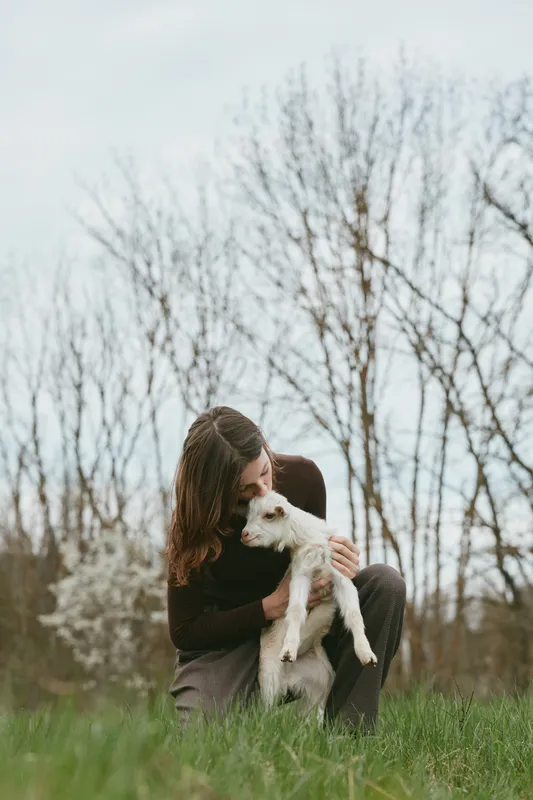 A woman outdoors holds and comforts a small goat kid, seated on green grass with bare trees in the background, creating a warm, intimate lifestyle moment focused on care and companionship.