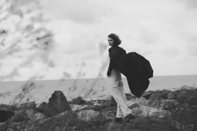 Woman on a rocky coast, flowing coat in wind, monochrome landscape, movement and solitude in an open wild scene