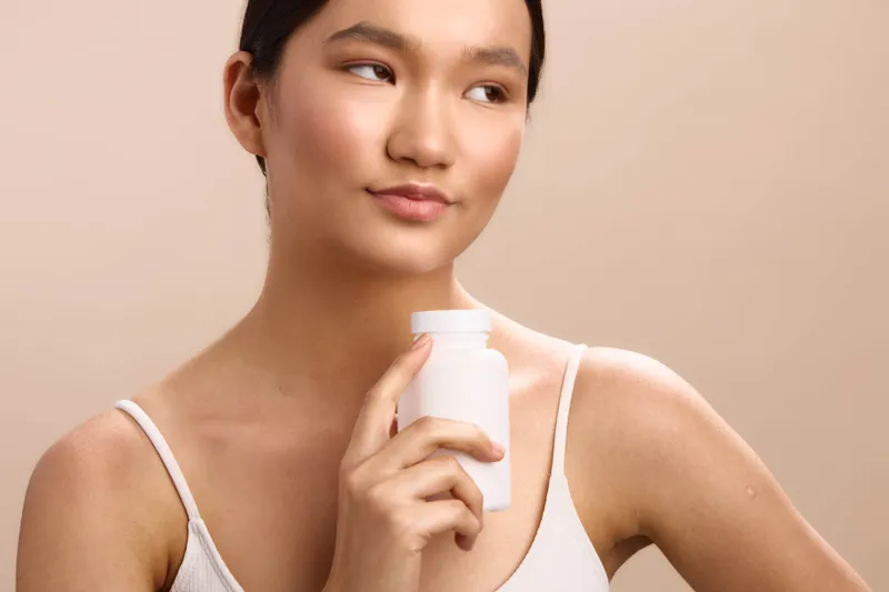 Asian woman holds a bottle against a clean studio backdrop, smiling softly as she contemplates skincare steps, creating a calm and fresh beauty atmosphere.