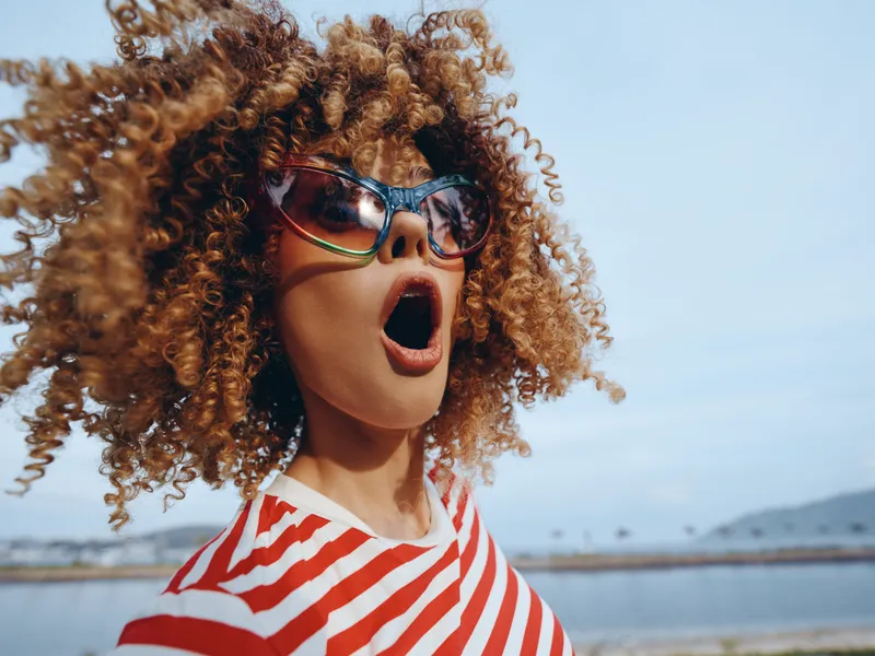 Vibrant portrait of a young woman with voluminous curly hair and bold sunglasses, mouth open in surprise, wearing a red and white striped shirt at the seaside.