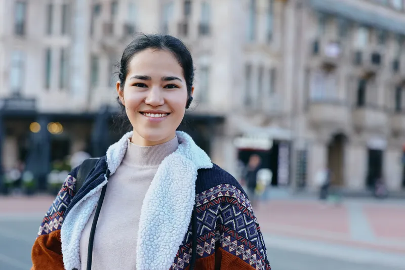 Smiling woman in a cozy layered outfit stands on a city street with urban architecture, showcasing street fashion, warmth and confidence in a daytime urban scene