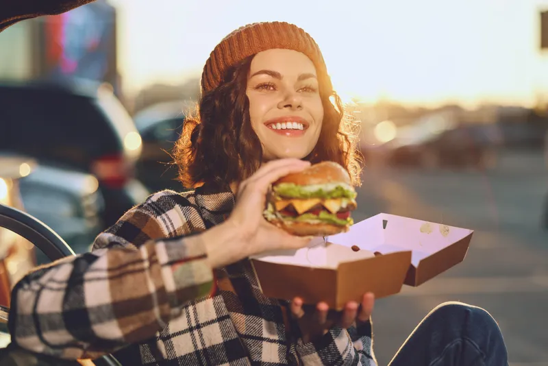 woman burger smile takeout parking sunset beanie plaid. Authenticity and candid lifestyle capture a joyful urban moment, golden hour glow on cozy meal and emotional storytelling in mindful living