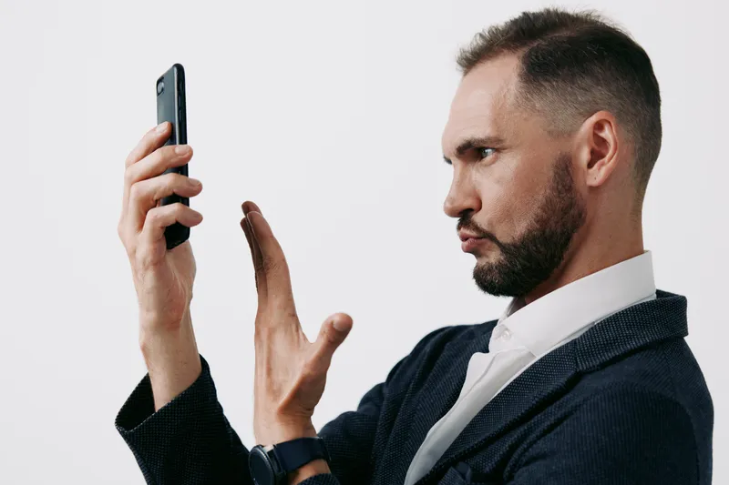 A professional business man stands isolated against a solid light background, with a focused posture, serious expression, and confident gesture as he speaks into a smartphone.