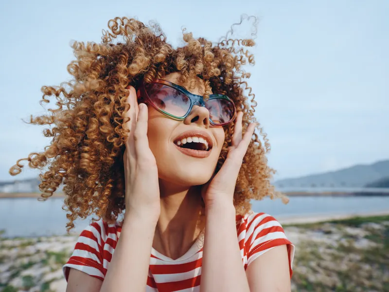 Joyful young girl with curly hair wearing sunglasses and a red striped shirt smiles outdoors at the beach, exuding carefree summer happiness and bright sunlight.