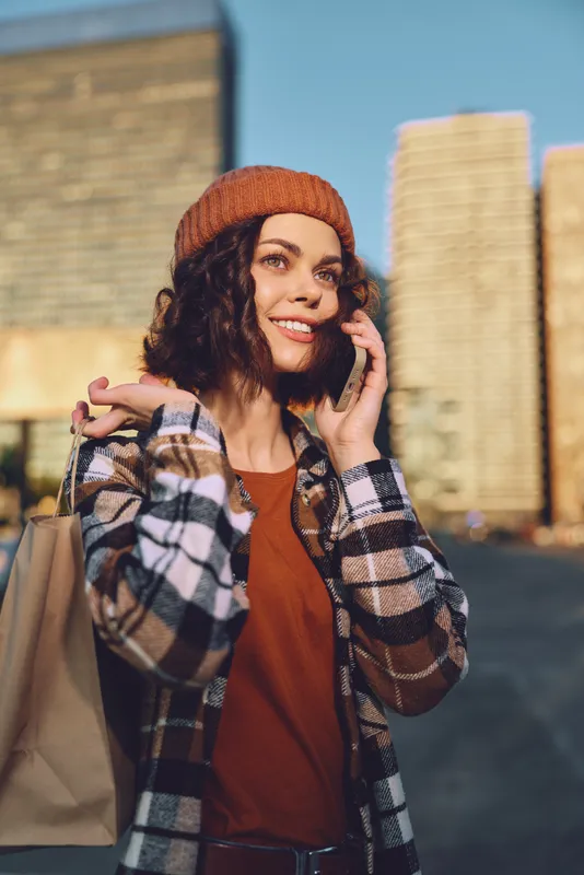 Woman shopping on city street, phone call and smiling in golden hour glow. Candid lifestyle shot conveying authenticity, mindful living and emotional storytelling in urban setting