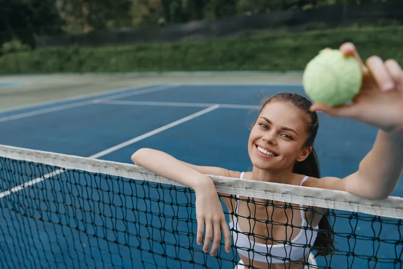 Joyful young woman holding a tennis ball, smiling at the camera on a blue tennis court, embodying an active lifestyle and sports enthusiasm