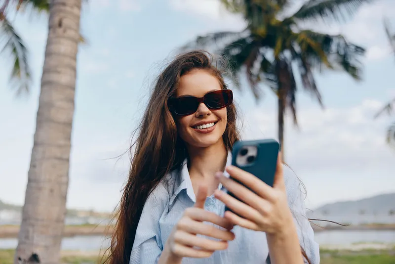 Улыбающаяся женщина в солнцезащитных очках держит мобильный телефон и позирует на фоне пальм