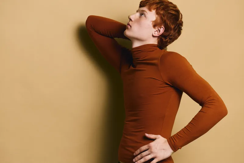 Redhead man posing in brown turtleneck sweater on beige background with hand on waist and looking upward, studio portrait with contemplative expression and casual style.