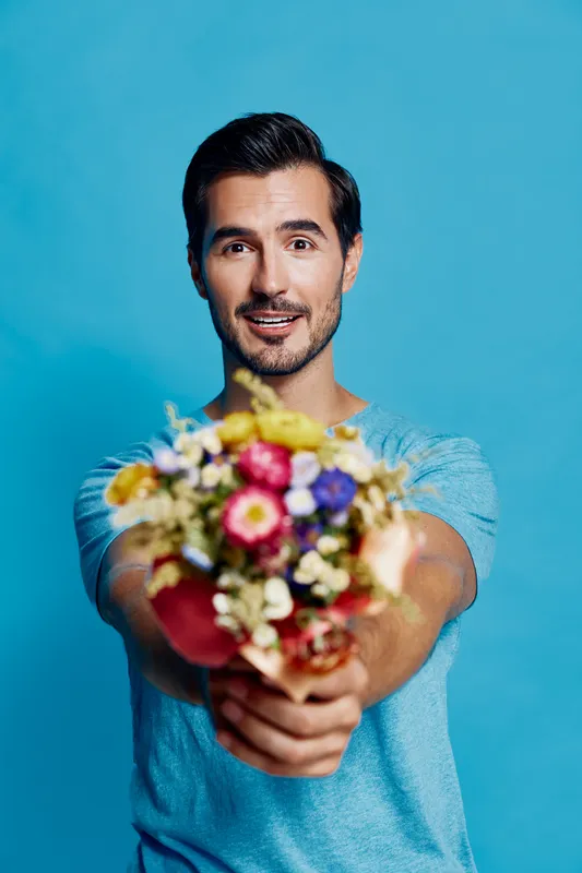 Smiling man holding colorful bouquet of flowers, wearing casual blue t-shirt, looking cheerful at the camera against a bright blue background. Flower gift concept.