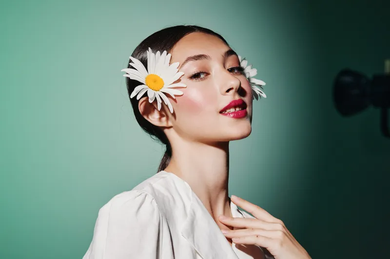 Young woman in white blouse with bright makeup wears white daisies by ears against mint background. Creative fashion beauty portrait highlighting flower accessories and glossy skin.