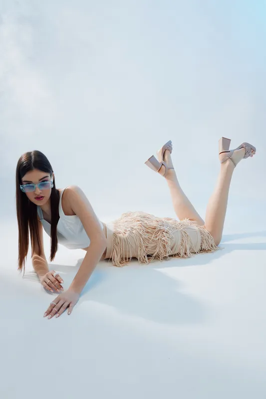 Young woman in white top and fringed skirt lies on white floor wearing sunglasses and heels. Fashion concept with beauty and style in bright studio light.