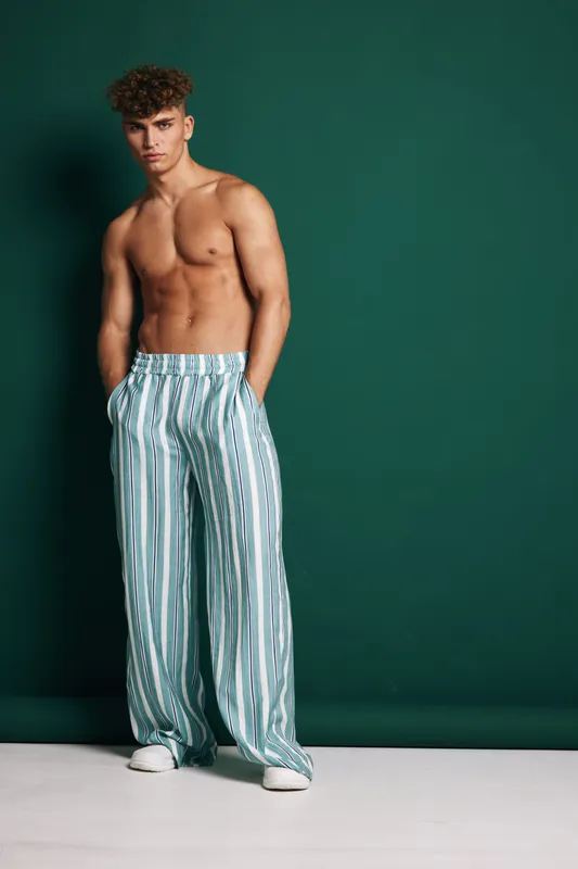 Young muscular man standing shirtless with hands in pockets wearing striped wide leg pants and white sneakers against green background in studio setting.