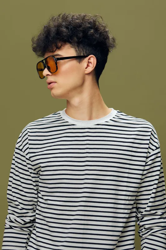 young man with curly hair wearing striped shirt and orange sunglasses looking sideways against olive green background, casual style portrait