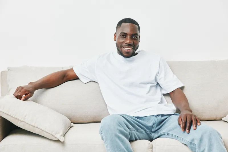 Cheerful young Black man in casual outfit smiling while sitting on a beige couch, showcasing relaxed home atmosphere and happiness