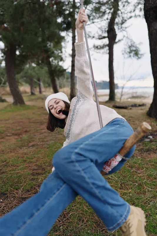 Girl swinging on a rope swing in a park among trees, wearing a beanie and sweater, enjoying carefree outdoor fun and laughter under soft daylight in a natural forested setting.