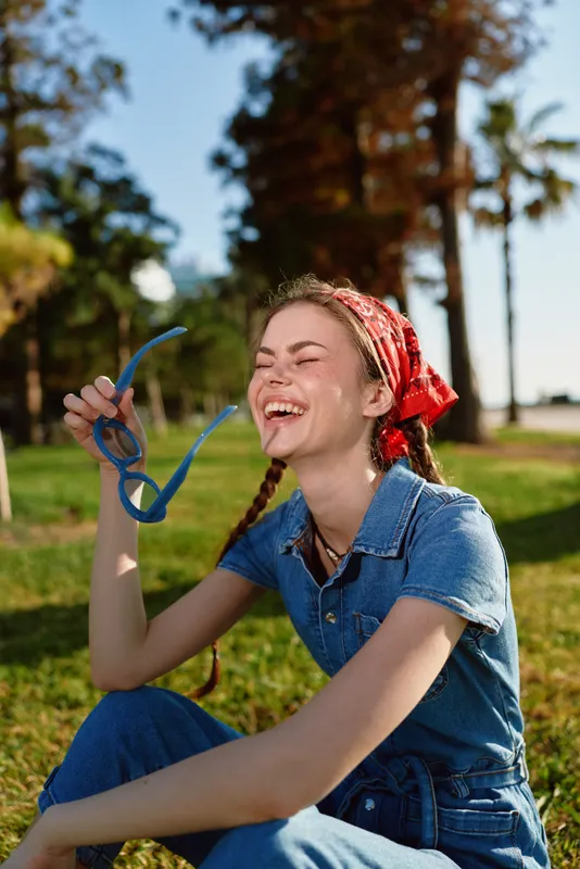 joyful woman in denim outfit laughing with sunglasses, wearing red bandana and boho-western street style, bright film color outdoors under sunny sky