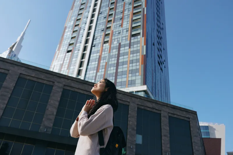 Woman gazing upward in an urban canyon of glass and steel, hands in prayer near a modern skyline, capturing hope, focus, and contemplation amid tall buildings and clear blue sky.