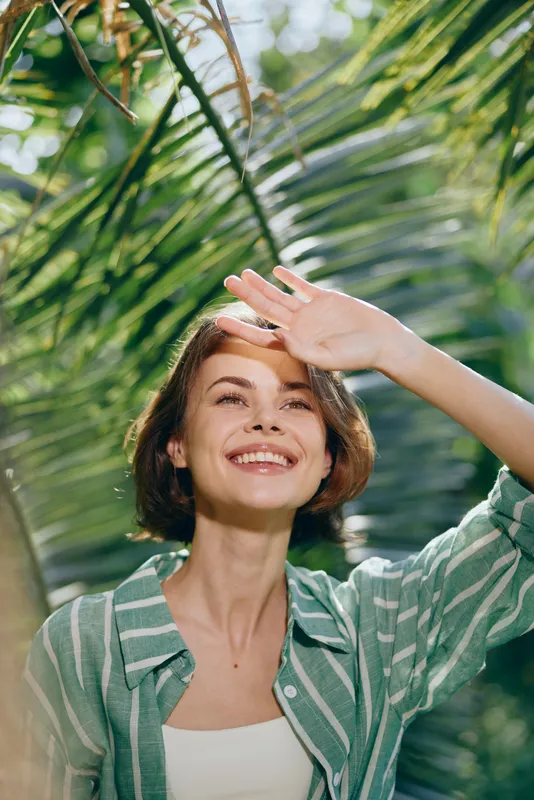 woman, smile, cheerful woman stands outdoors with sun in her hand shielding eyes, bright smile, lush greenery, warm light, natural portrait, carefree vibe, outdoor scene