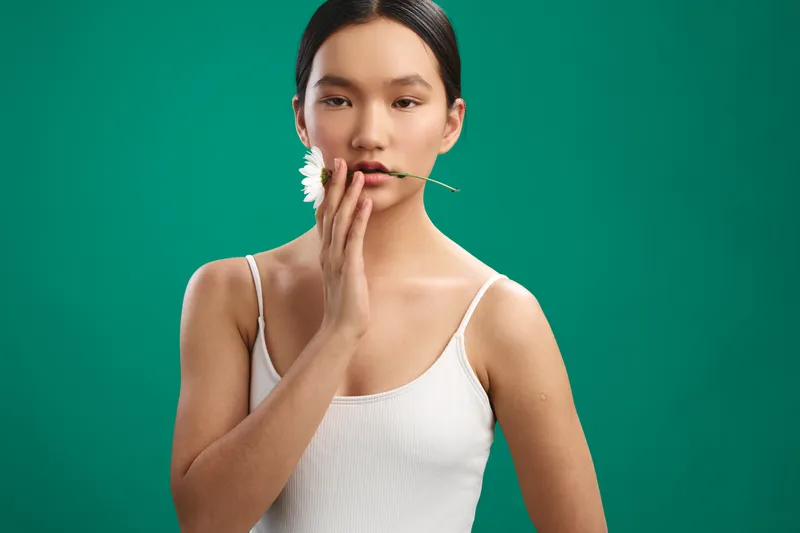 Asian woman with a delicate smile stands in a clean studio setting, gently touching a flower near her lips, conveying calm confidence and artistic mood.