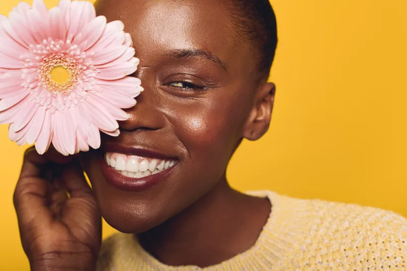 Close-up of a smiling dark skinned African woman holding a pink flower near her face against a yellow background. Concept of natural beauty, happiness, skincare, and floral freshness.