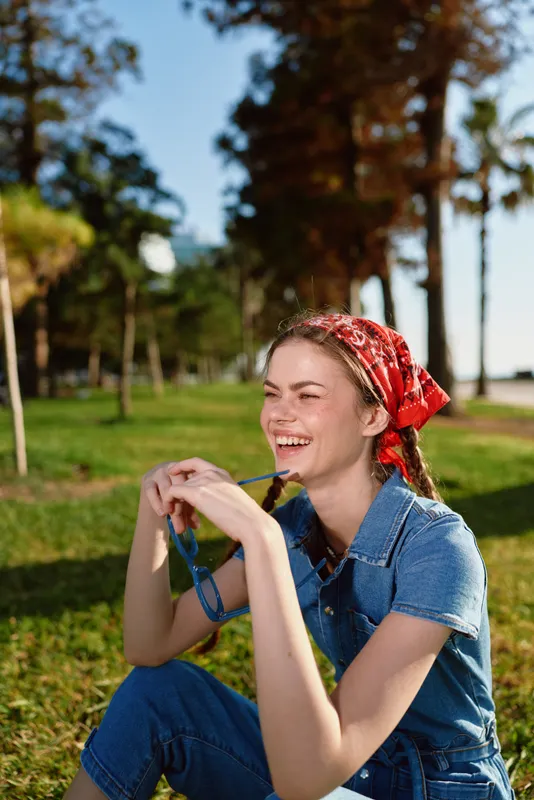 lifestyle woman in modern boho-western denim street style sitting outdoors smiling with red bandana and braided hair, film color tone, vibrant natural park background